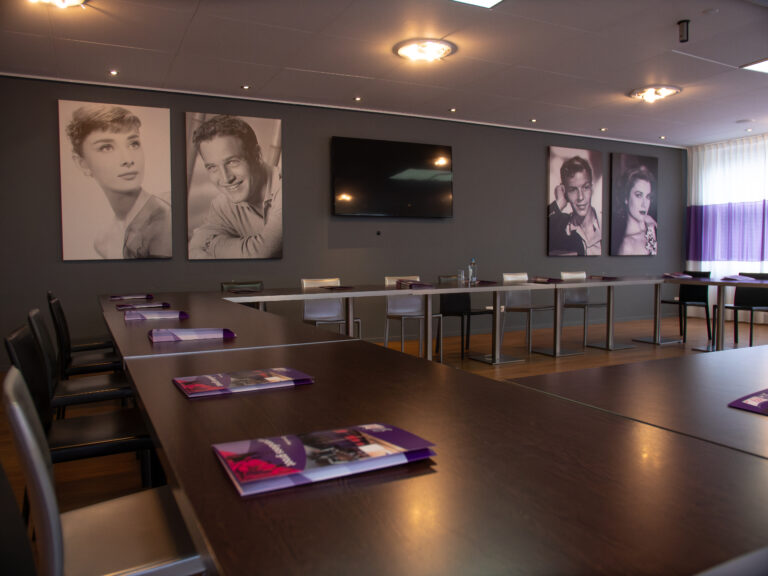 Moderne vergaderruimte met U-vormige tafels, tv-scherm en vier zwart-wit filmsterrenportretten met paarse details.