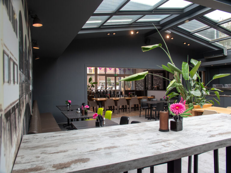 Licht, modern restaurantinterieur met dakramen, donkere muren, diverse tafels en groene planten.