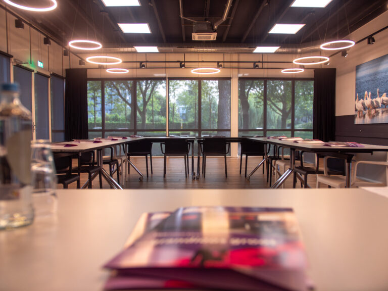 Ruime, moderne conferentiezaal met U-opstelling, ringverlichting, uitzicht op bomen en brochures vooraan.