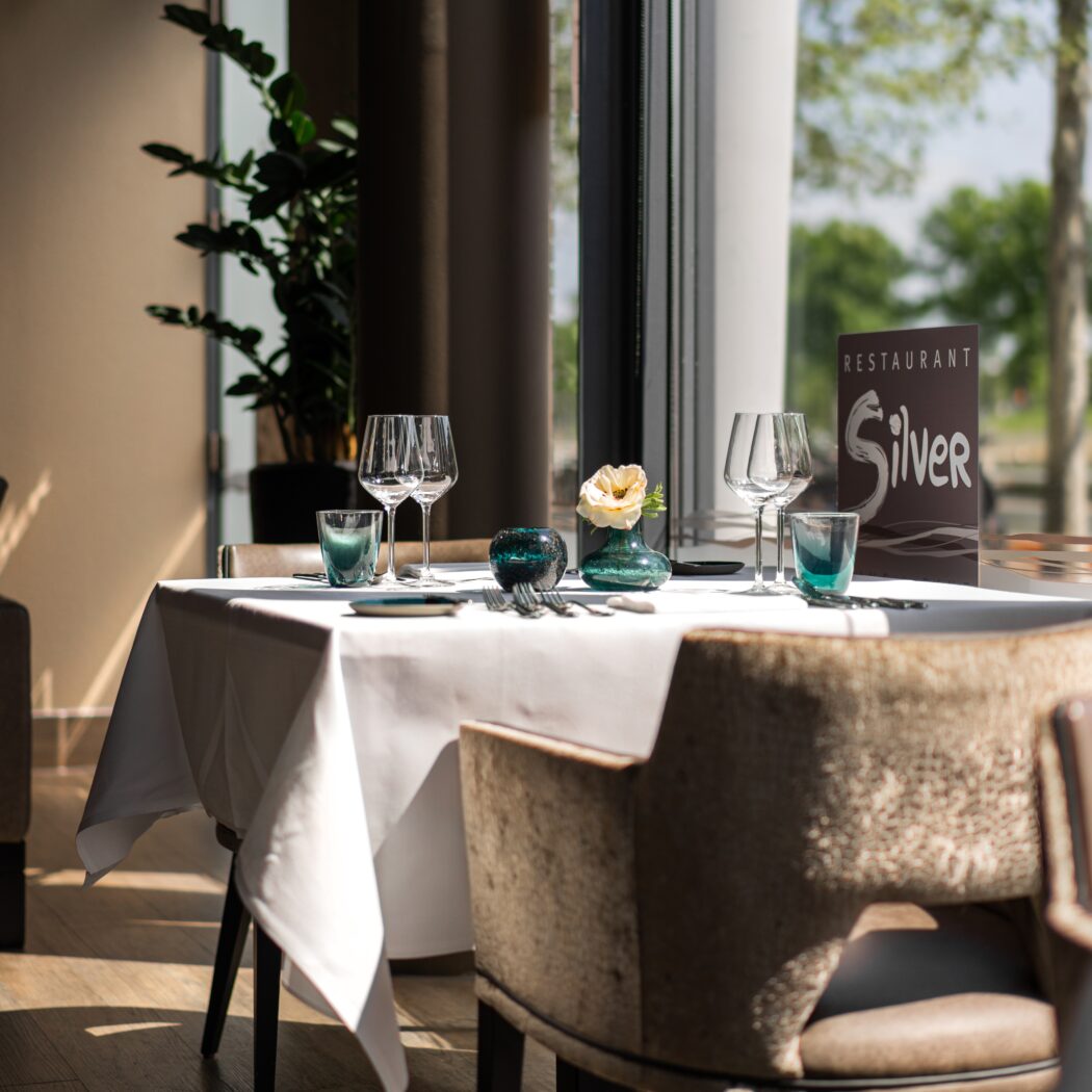 Gedekte tafel in Restaurant Silver met witte linnen, turquoise glazen, bloem en zonnig uitzicht.