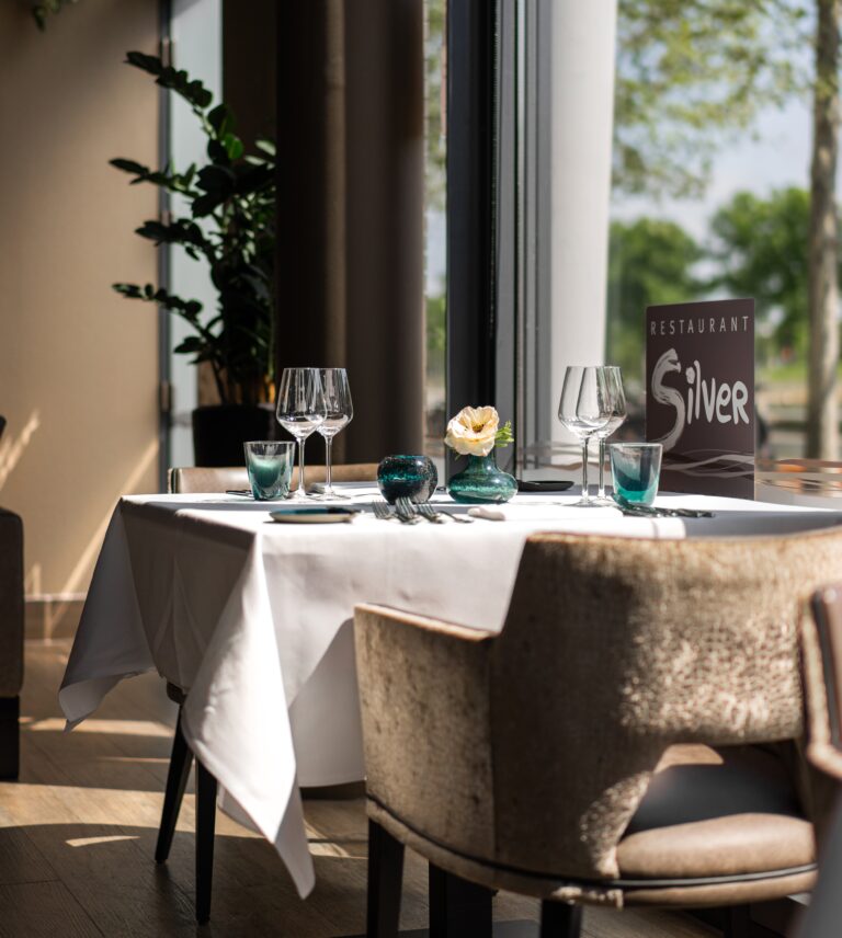 Gedekte tafel in Restaurant Silver met witte linnen, turquoise glazen, bloem en zonnig uitzicht.