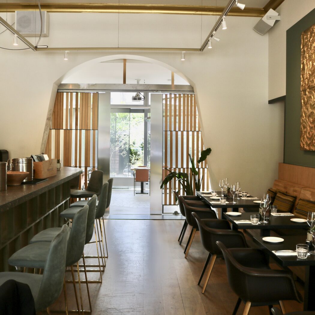 Sfeervol restaurantinterieur met een lange bar, grijze barkrukken, gedekte eettafels en een opvallend gouden wandkunstwerk.