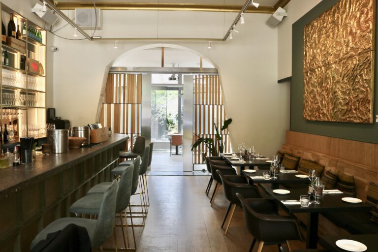 Sfeervol restaurantinterieur met een lange bar, grijze barkrukken, gedekte eettafels en een opvallend gouden wandkunstwerk.