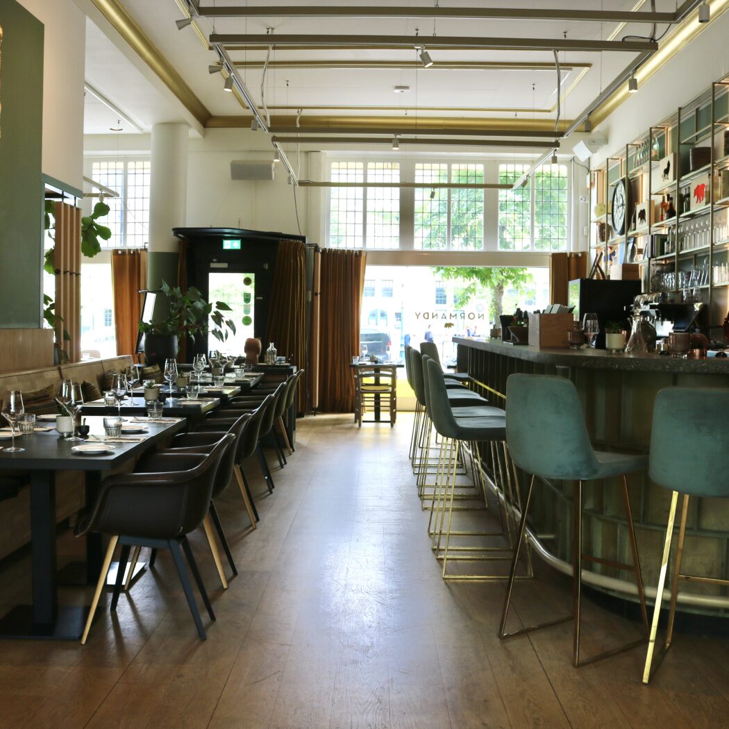 Stijlvol restaurantinterieur met lange eettafel, bar met teal krukken, een gouden kunstwerk en NORMANDY reflectie.