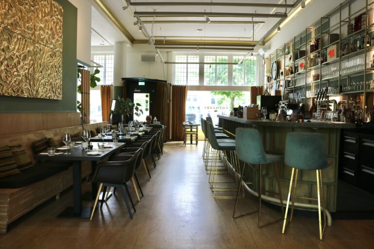 Stijlvol restaurantinterieur met lange eettafel, bar met teal krukken, een gouden kunstwerk en NORMANDY reflectie.