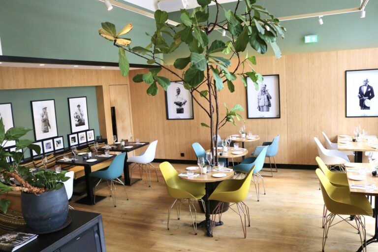 Stijlvol restaurant met houten wanden, mintgroene accenten, ingedekte tafels, kleurrijke designstoelen en grote ficus.