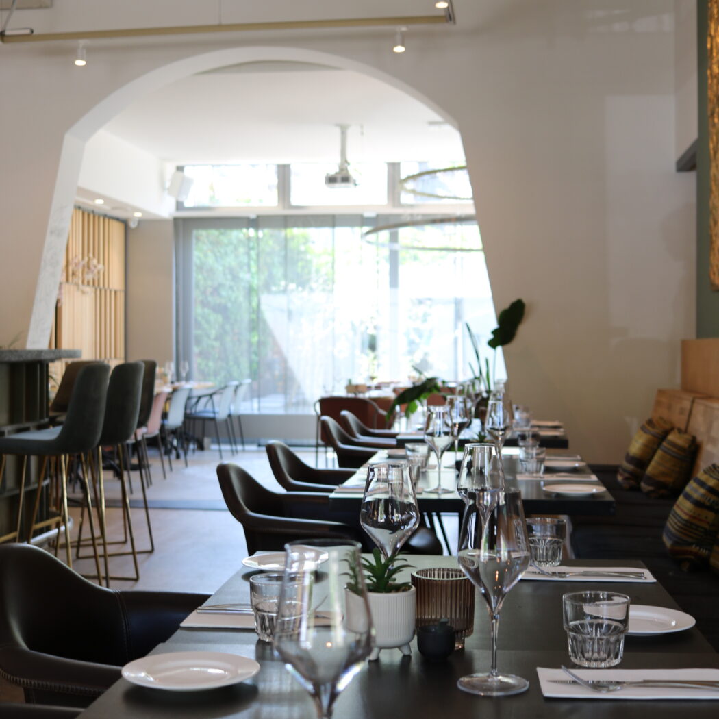 Modern restaurantinterieur met gedekte tafels en wijnglazen, leidend naar een lichte eetruimte achter een boog.