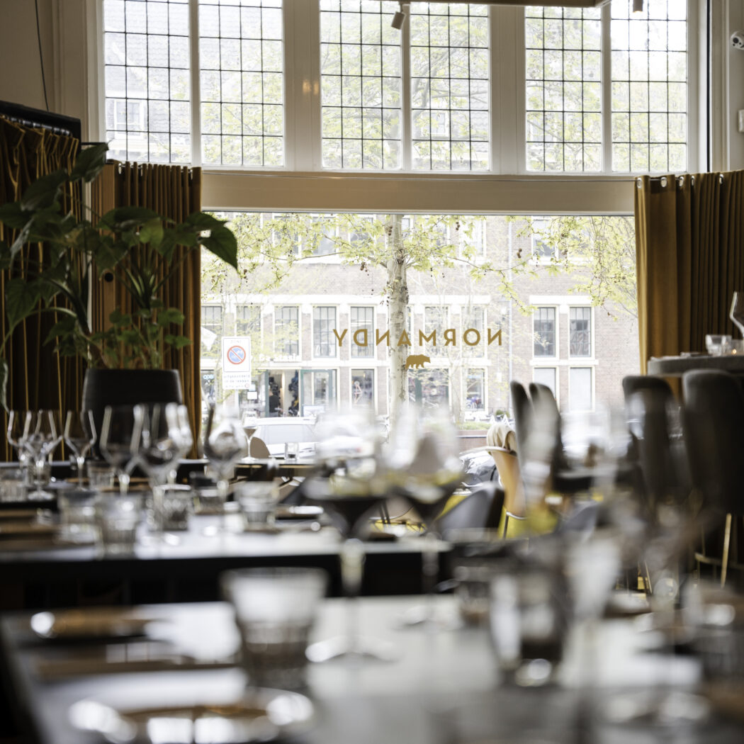 Interieur van een restaurant met gedekte tafels, grote ramen en het NORMANDY logo met beer.