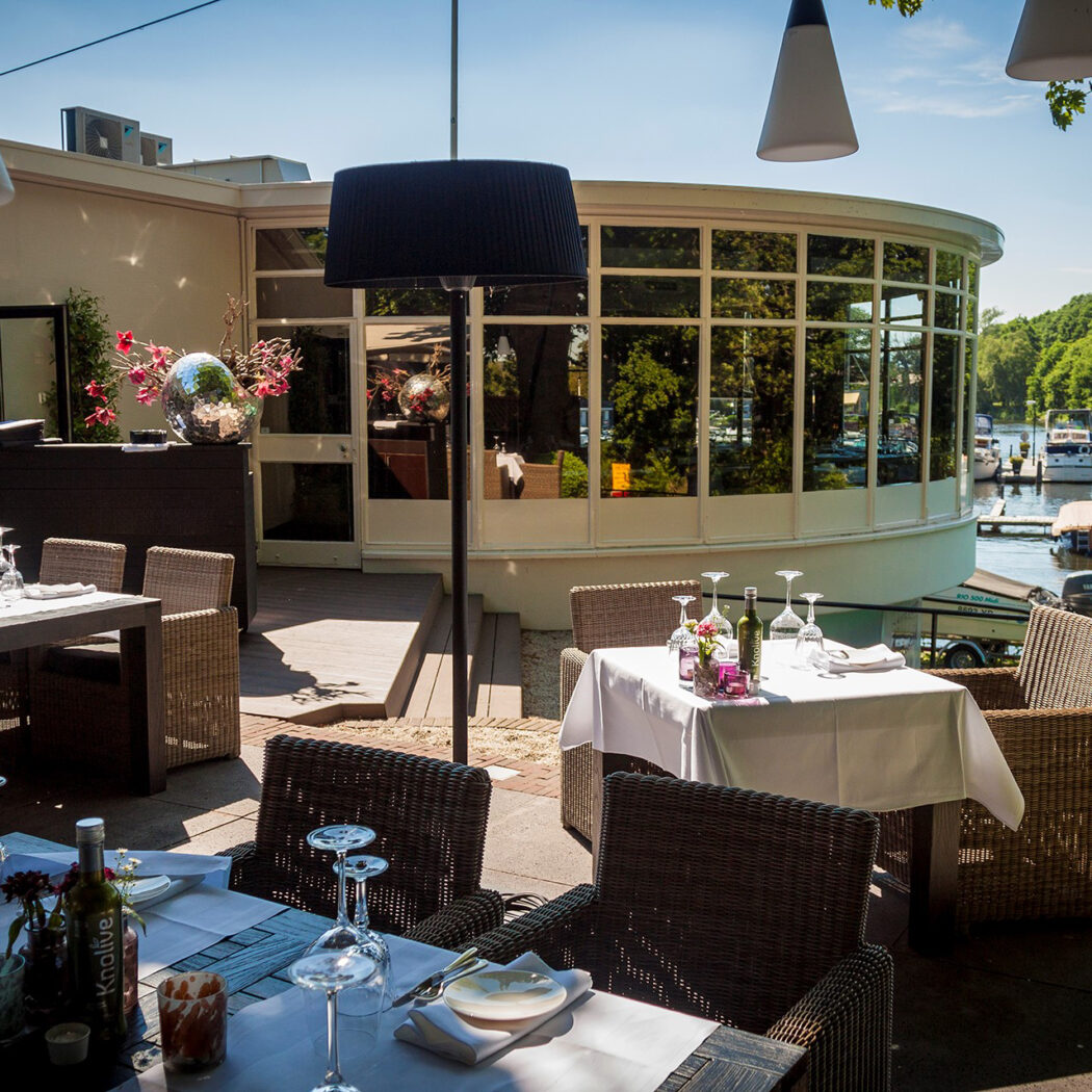 Zonnig restaurantterras met gedekte tafels en rotan stoelen, uitkijkend op een waterweg met jachthaven.