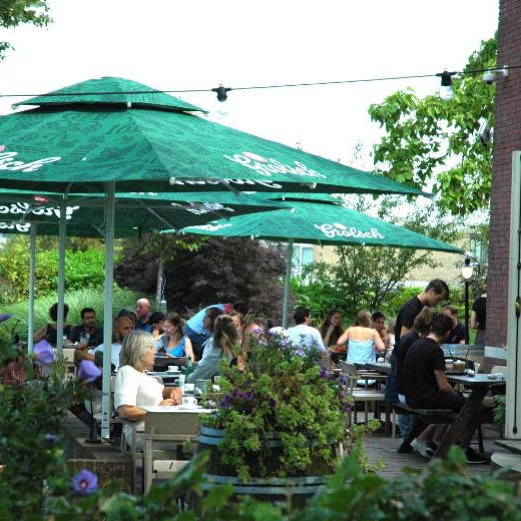 Druk terras met gasten die genieten onder groene Grolsch parasols, omringd door weelderige planten.