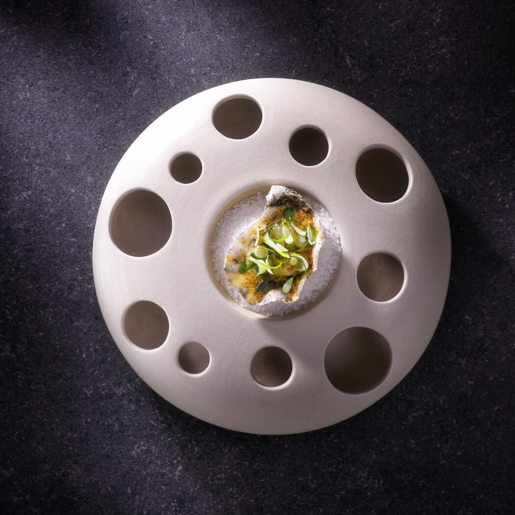 Oester met groene garnering en saus op een zoutbedje, geserveerd op een wit keramisch bord met ronde gaten.