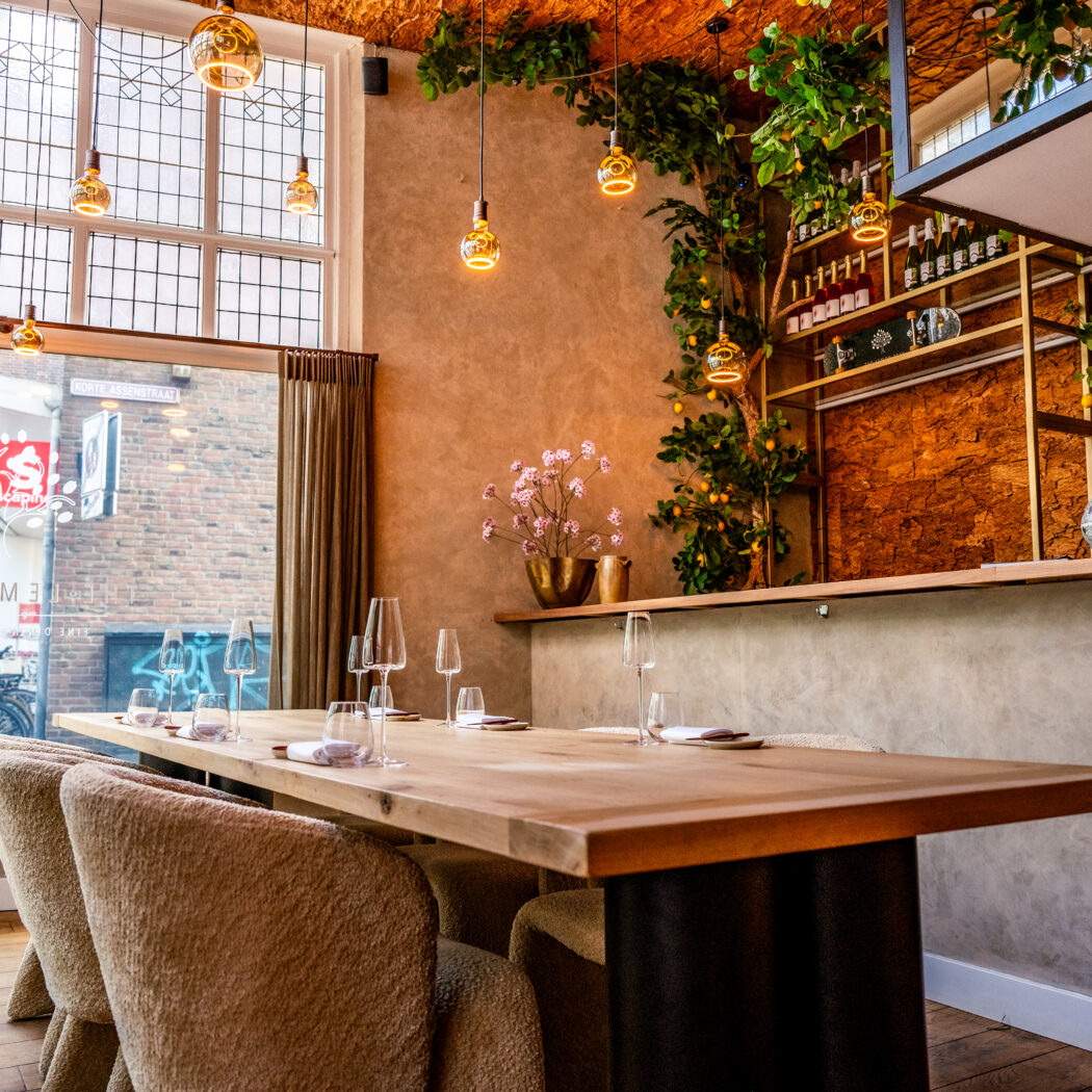 Sfeervol restaurant interieur met lange houten eettafel, pluche stoelen en botanische decoratie. De naam LEMON is zichtbaar op het raam.