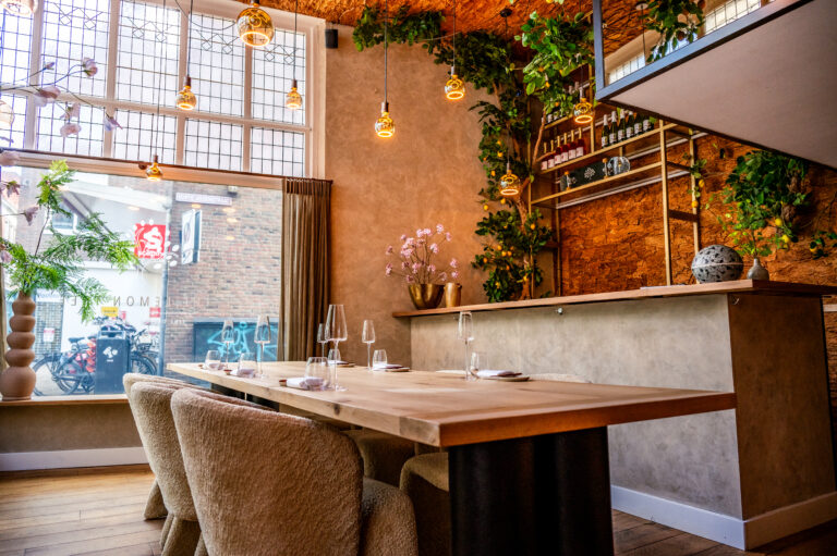 Sfeervol restaurant interieur met lange houten eettafel, pluche stoelen en botanische decoratie. De naam LEMON is zichtbaar op het raam.
