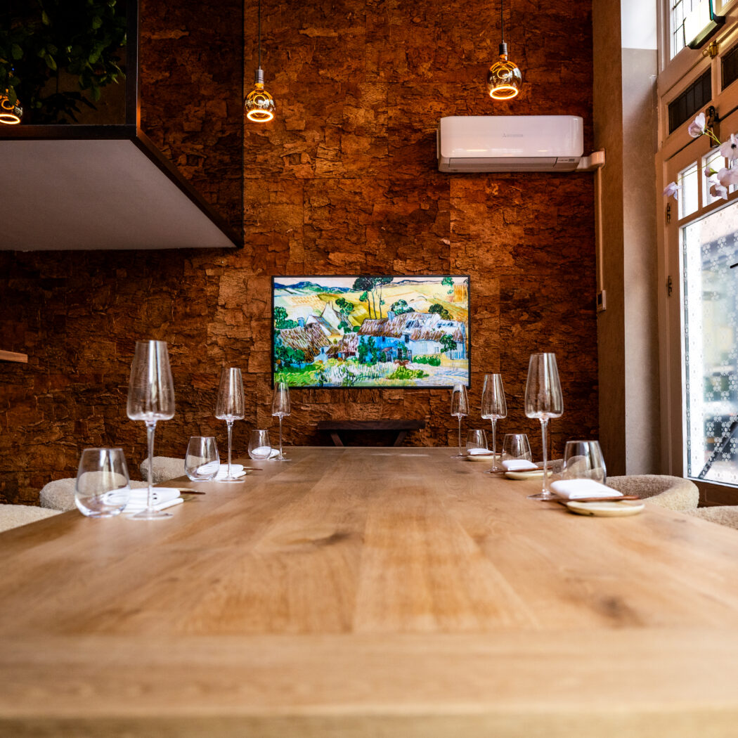 Lange houten eettafel met elegante wijnglazen en servies, voor een gestructureerde muur met kleurrijk schilderij.