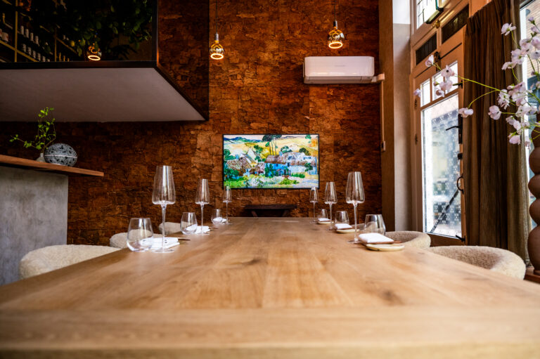 Lange houten eettafel met elegante wijnglazen en servies, voor een gestructureerde muur met kleurrijk schilderij.