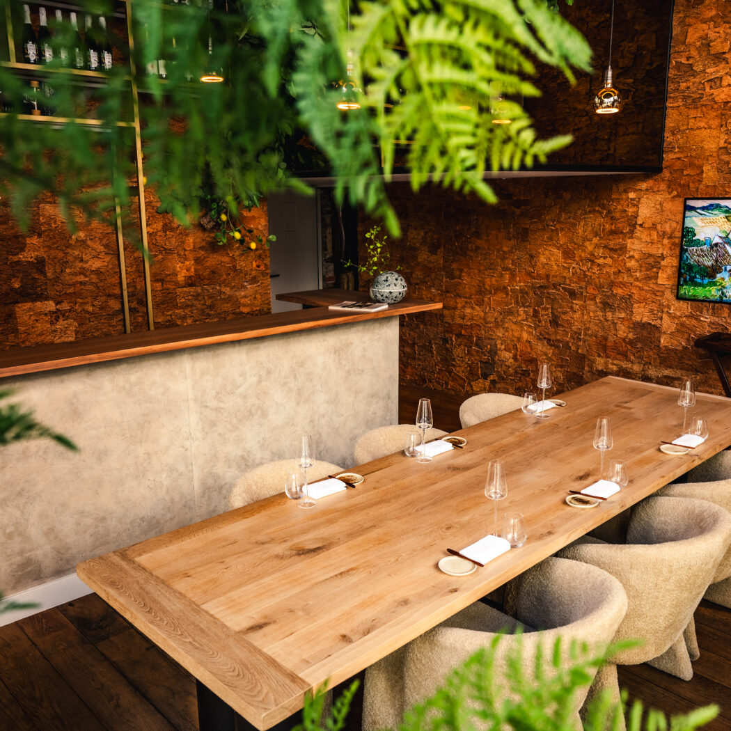 Lange houten eettafel met gedekte plaatsen en beige stoelen in een sfeervol restaurant met bakstenen muur en planten.