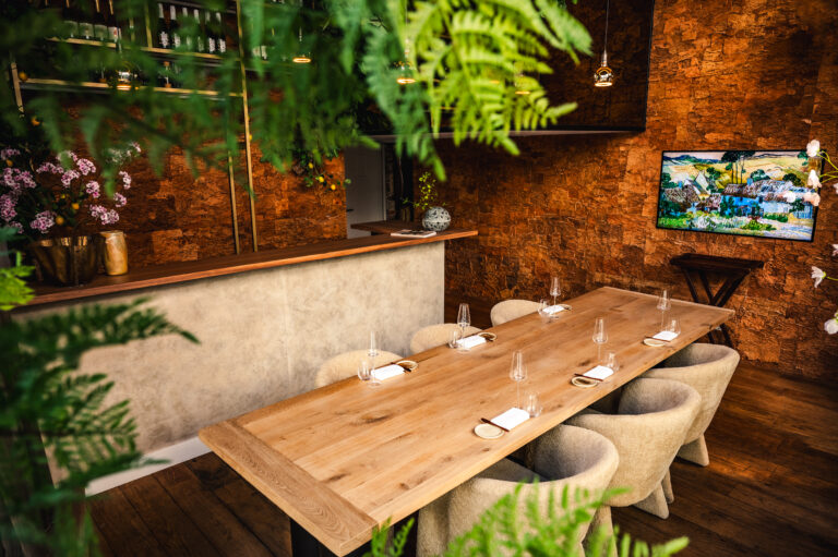 Lange houten eettafel met gedekte plaatsen en beige stoelen in een sfeervol restaurant met bakstenen muur en planten.
