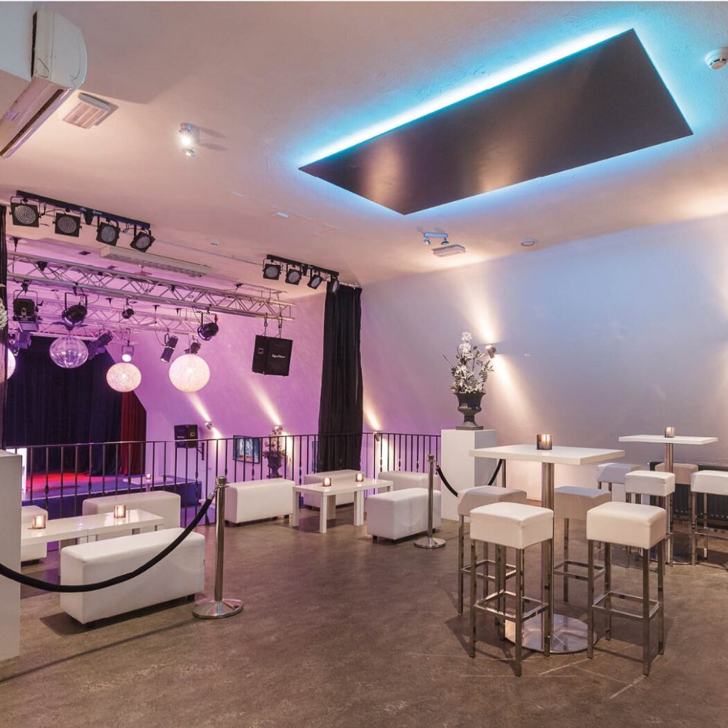 Moderne evenementenlocatie met podium, loungebanken, hoge tafels en sfeervolle verlichting in paars en blauw.