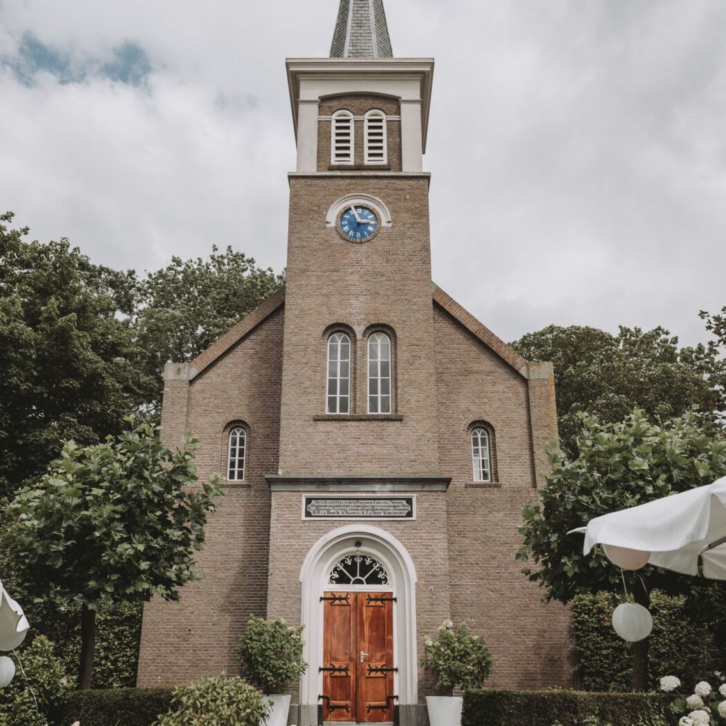Feestelijke bakstenen kerk met spitse toren, blauwe klok en zwarte loper naar houten deuren.