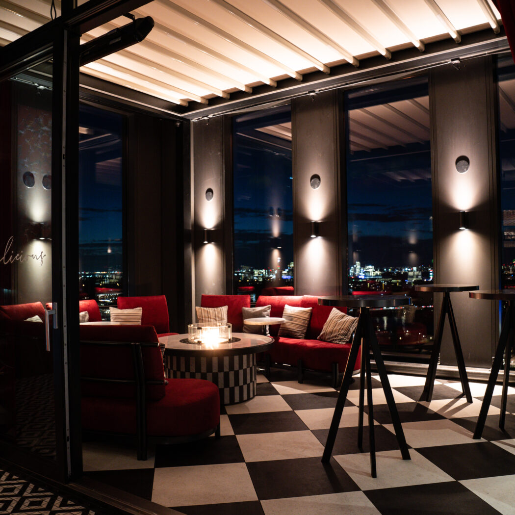 Sfeervolle rooftopbar met rode banken, vuurtafel, zwart-wit geruite vloer en Delicious neontekst met stadsgezicht.