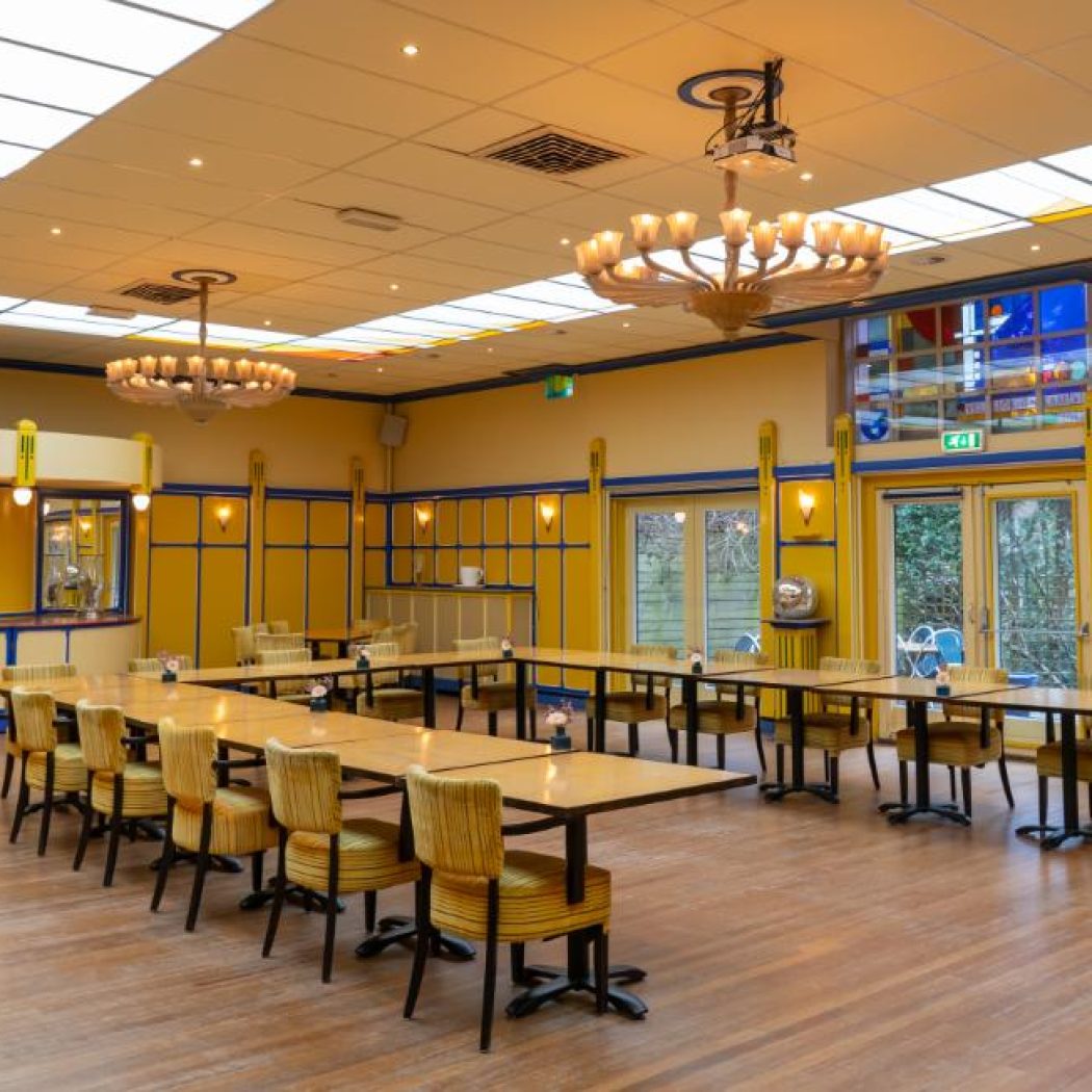 Ruime gele vergaderzaal met blauwe accenten, lange tafels, houten vloer en kleurrijke lichtpanelen.