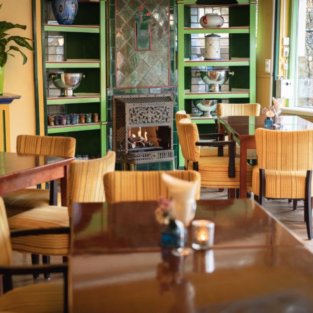 Gezellig restaurantinterieur met houten tafels, gele gestreepte stoelen, brandende haard en groene kasten.