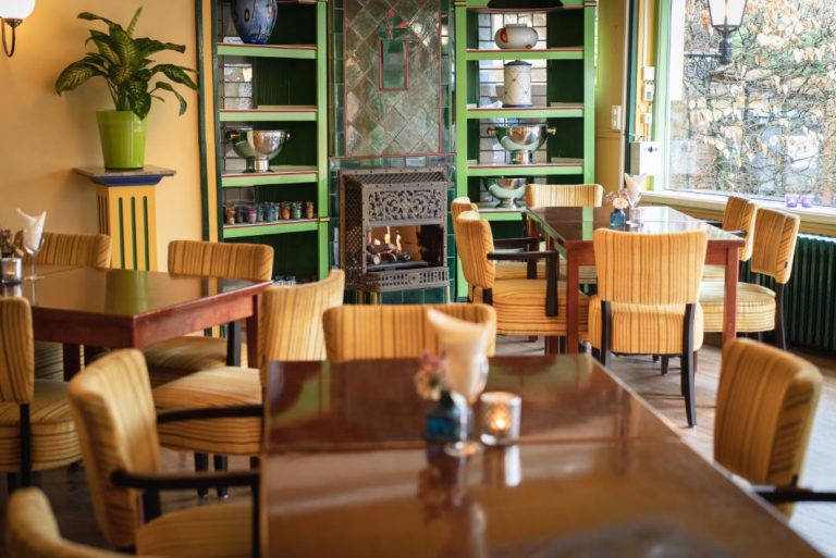 Gezellig restaurantinterieur met houten tafels, gele gestreepte stoelen, brandende haard en groene kasten.