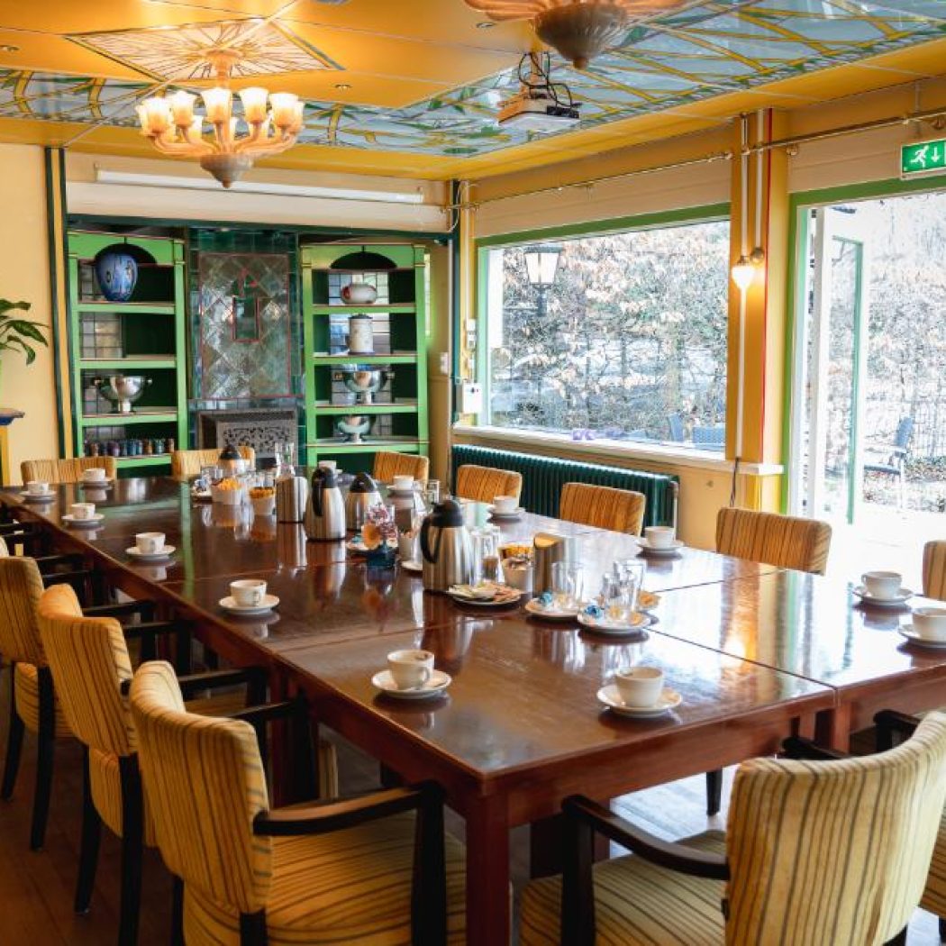 Sfeervolle vergaderzaal met decoratief plafond, lange houten tafel met koffieservies, groene kasten en winterzicht.