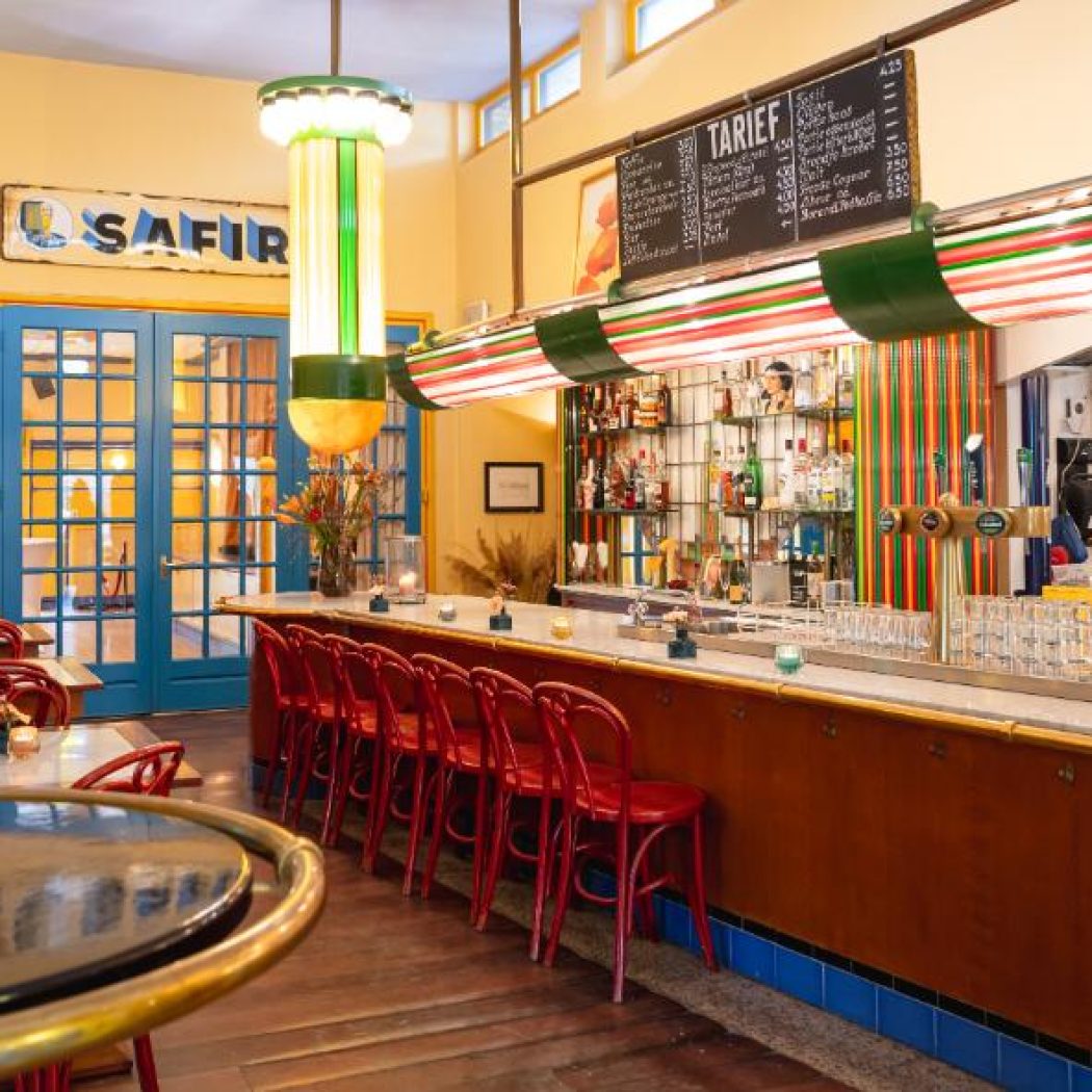 Gezellige vintage bar met houten toog, rode krukken, gele muren en blauwe deuren met SAFIR logo.