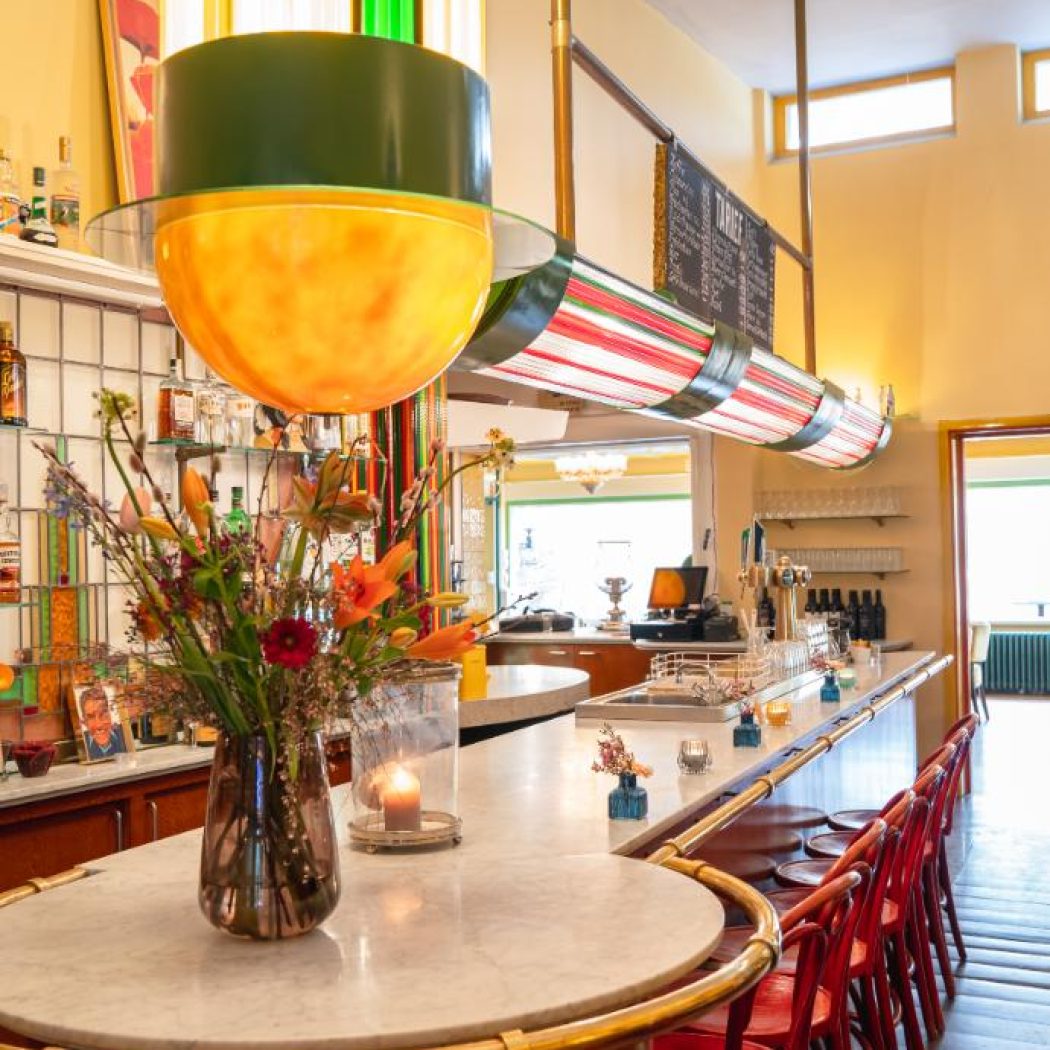 Stijlvol café interieur met marmeren bar, rode stoelen, bloemen en unieke gele en groene hanglampen.