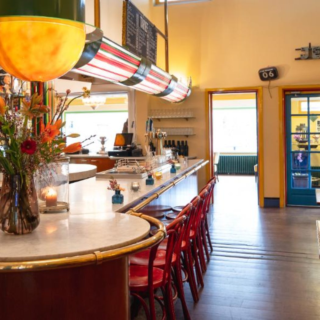Gezellig café interieur met lange marmeren bar, rode barkrukken, bloemen en een JENEVER bord boven blauwe deuren.