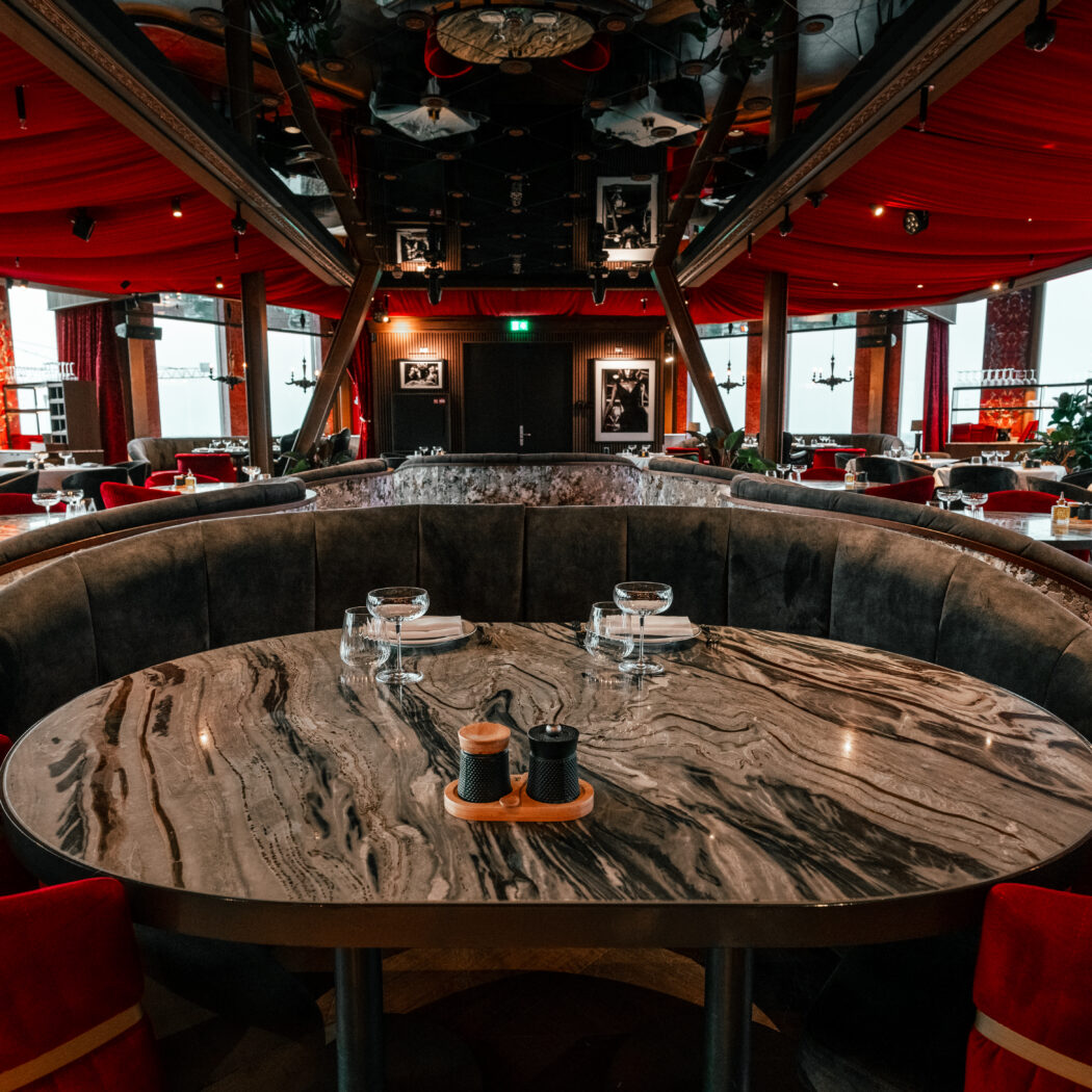 Luxe restaurantinterieur met rood fluwelen plafond, grijze ronde banken en marmeren tafels met rode stoelen.