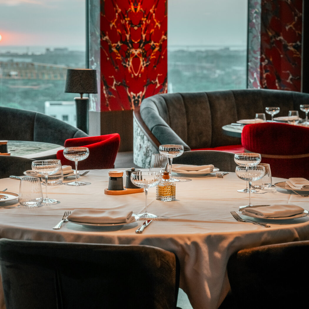 Elegante restaurantzaal met een rond gedekte tafel, donkere stoelen, en een panoramisch uitzicht op zonsondergang.