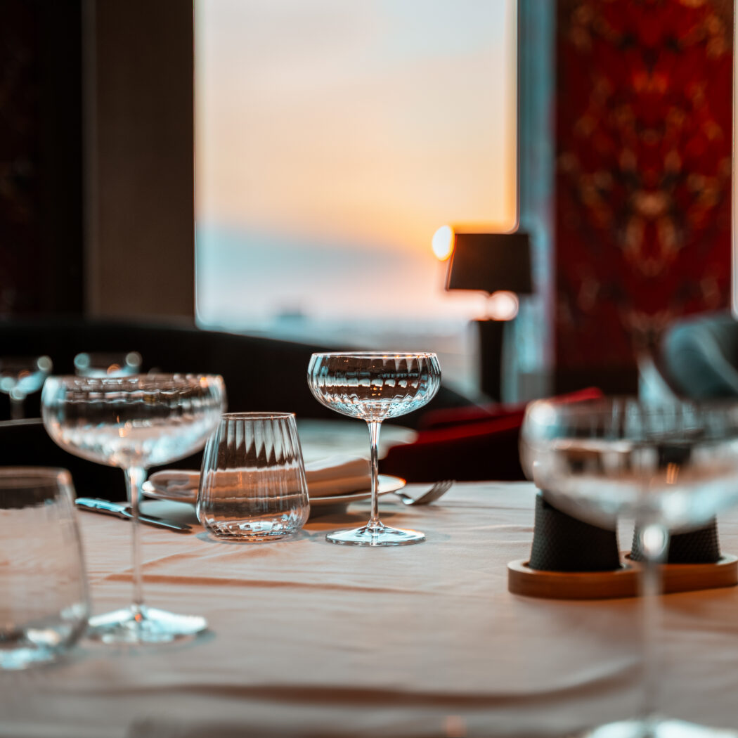 Gedekte tafel in een restaurant met diverse glazen, bestek en witte servetten, met een warme zonsondergang door het raam.