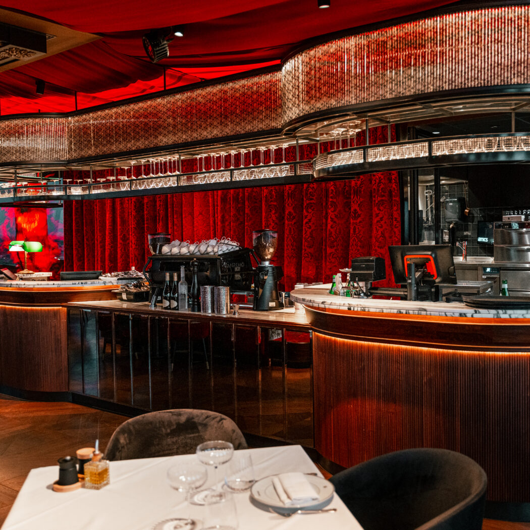 Verlichte bar met glaswerk en koffiezetapparatuur in een chic restaurant met rode fluwelen gordijnen.