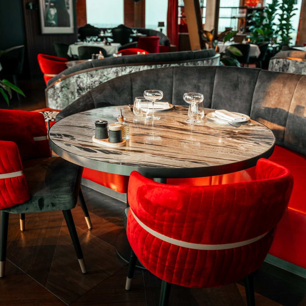 Ronde marmeren eettafel met rode fluwelen stoelen en een grijze gebogen bank in een elegant restaurant.
