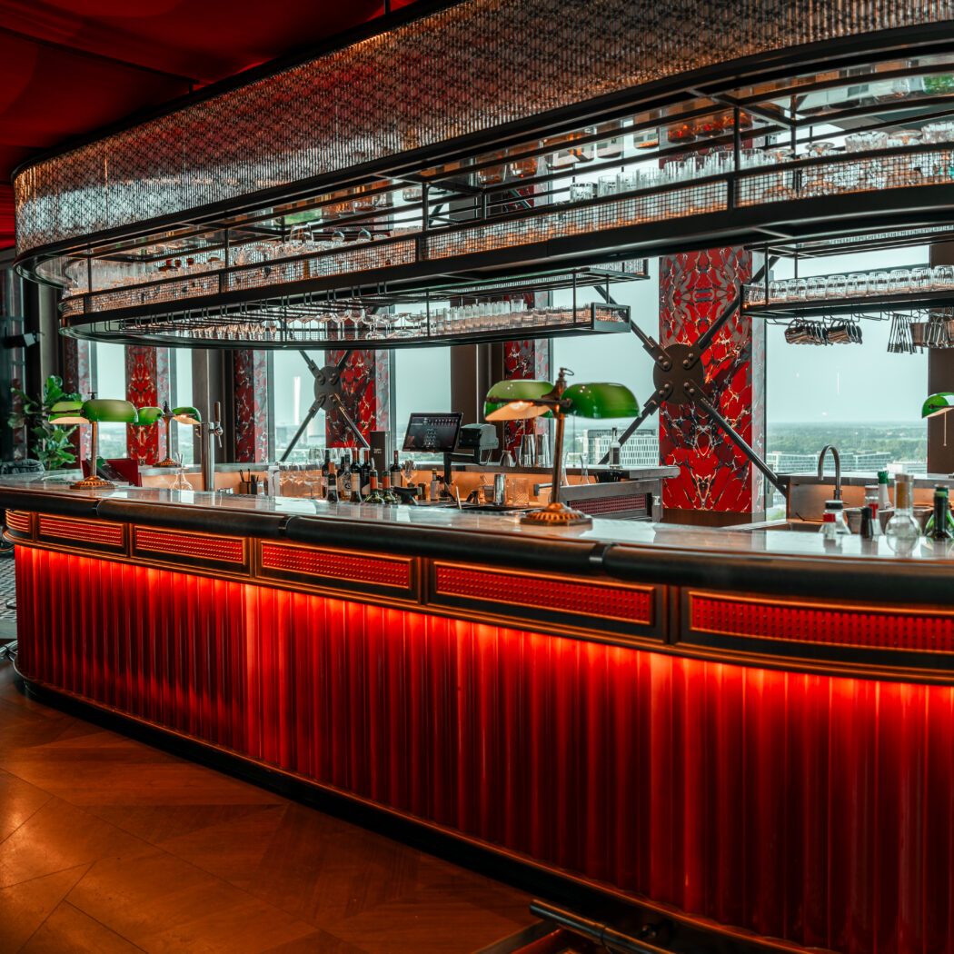 Een luxe, moderne cocktailbar met rood verlichte balie, groene lampen en uitzicht op de stad.