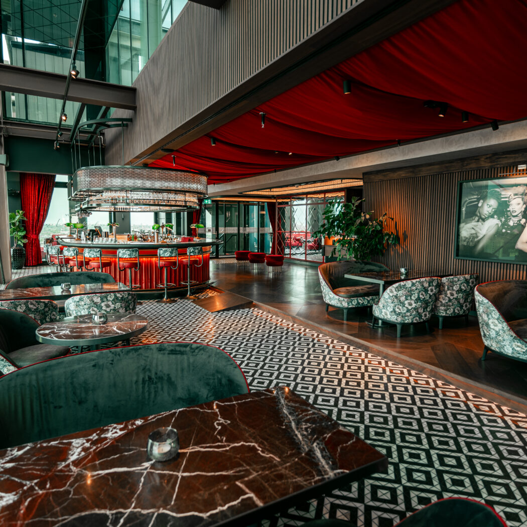 Luxe loungebar met rode bar, donkergroene zitmeubels, marmeren tafels, patroonvloer en rood gedrapeerd plafond.