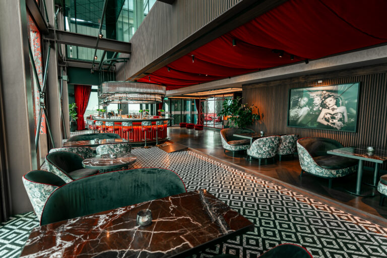Luxe loungebar met rode bar, donkergroene zitmeubels, marmeren tafels, patroonvloer en rood gedrapeerd plafond.