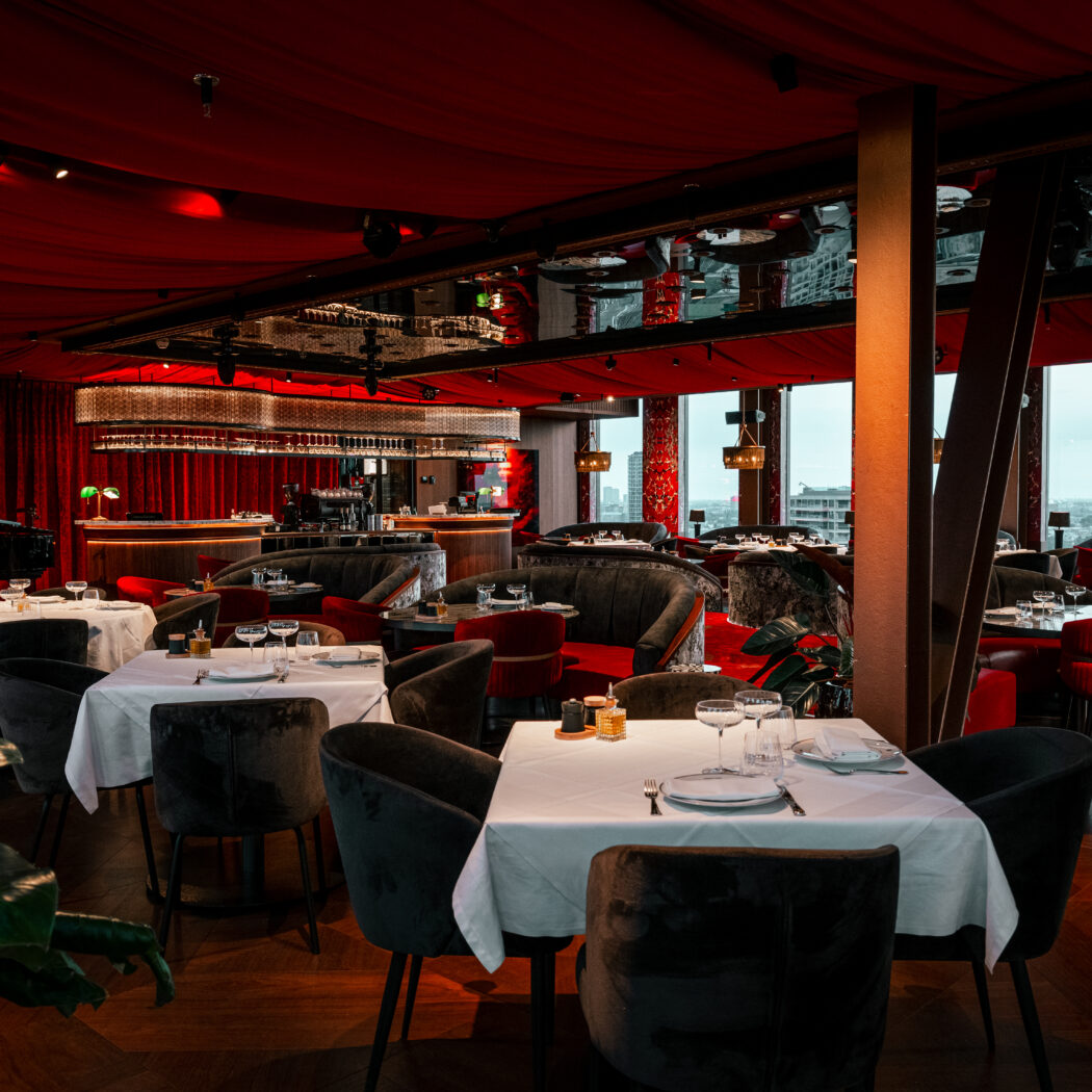 Stijlvol restaurant met rode plafonddraperieën, fluwelen meubels, gedekte tafels, een bar, vleugel en stadszicht.