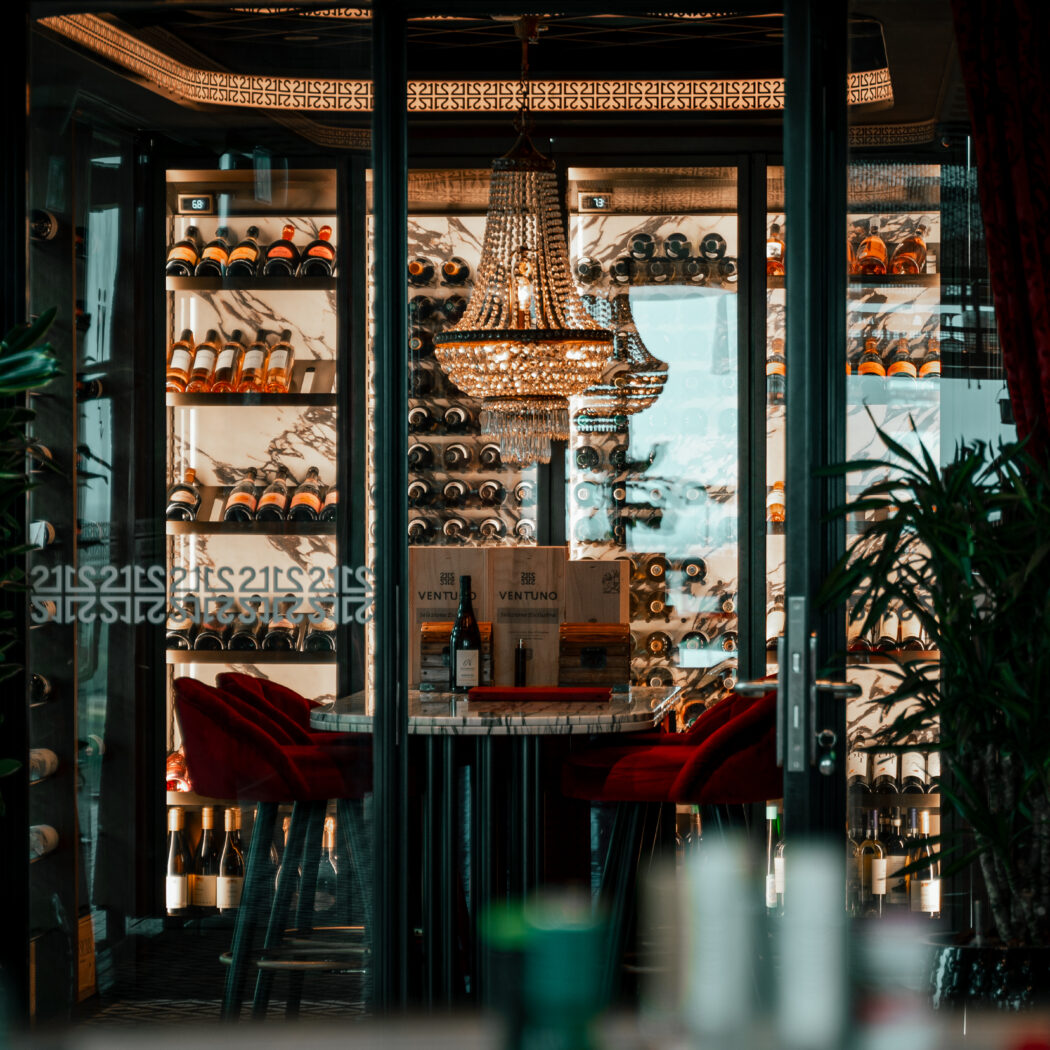 Luxe glazen wijnvitrine met flessen, kristallen kroonluchter, marmeren tafel met VENTUNO kist en rode fluwelen stoelen.