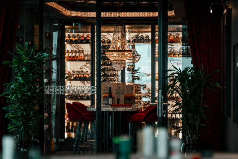 Luxe glazen wijnvitrine met flessen, kristallen kroonluchter, marmeren tafel met VENTUNO kist en rode fluwelen stoelen.