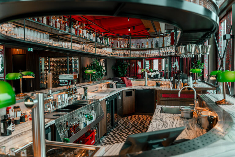 Luxe barinterieur met marmeren balie, gevulde schappen, groene bureaulampen en een rood accentplafond.