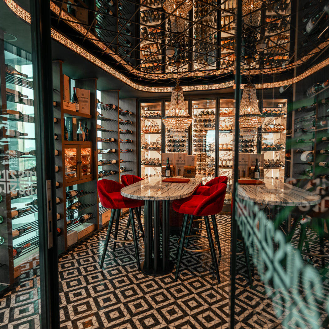 Elegante wijnkelder met glazen vitrines vol flessen, twee marmeren tafels, rode fluwelen barstoelen en kroonluchters.