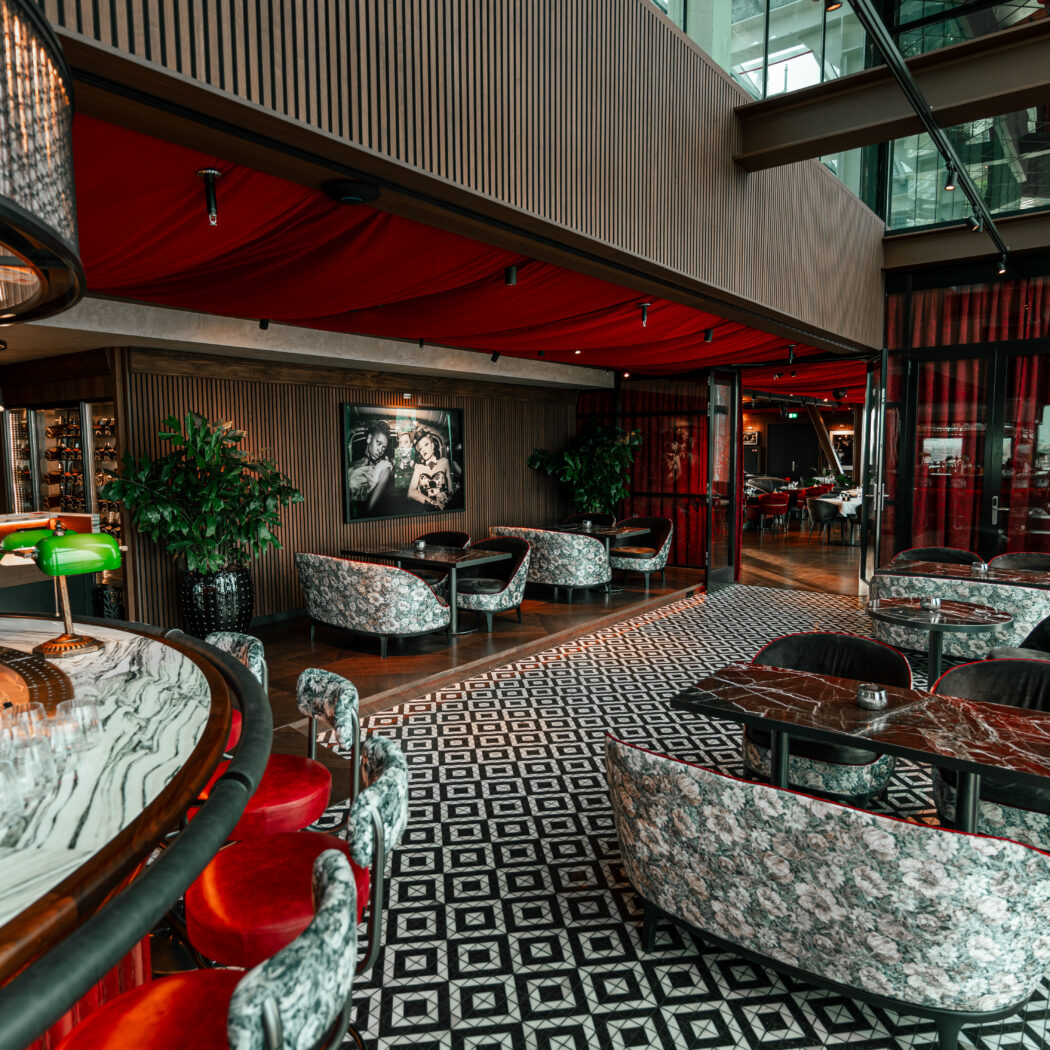 Luxe bar en lounge met rode draperieën, marmeren bar, en zitjes op een zwart-wit geometrische vloer.