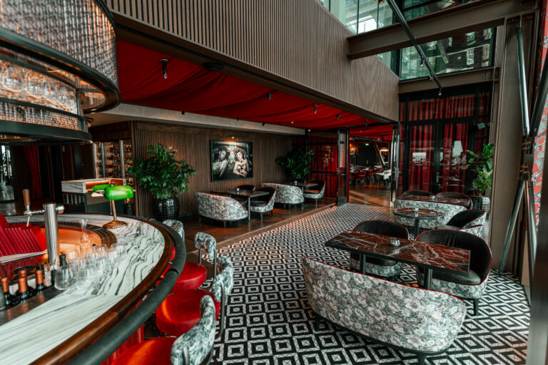 Luxe bar en lounge met rode draperieën, marmeren bar, en zitjes op een zwart-wit geometrische vloer.