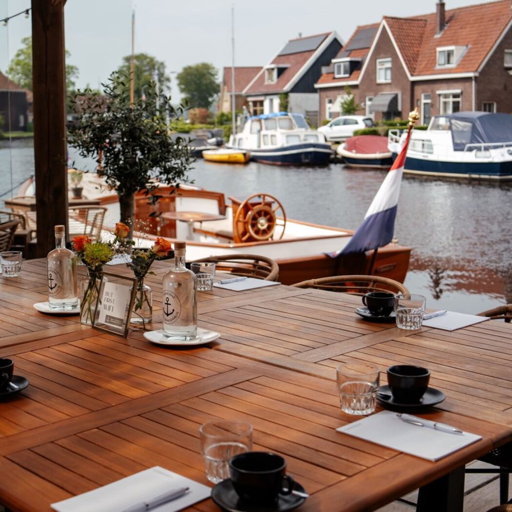 Grote houten vergadertafel op een terras aan de gracht met boten, huizen en een BUT FIRST WIFI bordje.