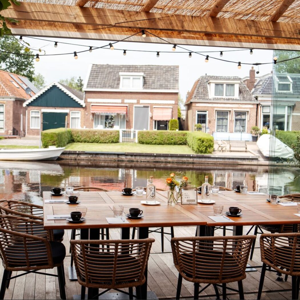 Sfeervol restaurantterras aan een gracht met gedekte houten tafels, rieten stoelen en een pergola met lampjes.