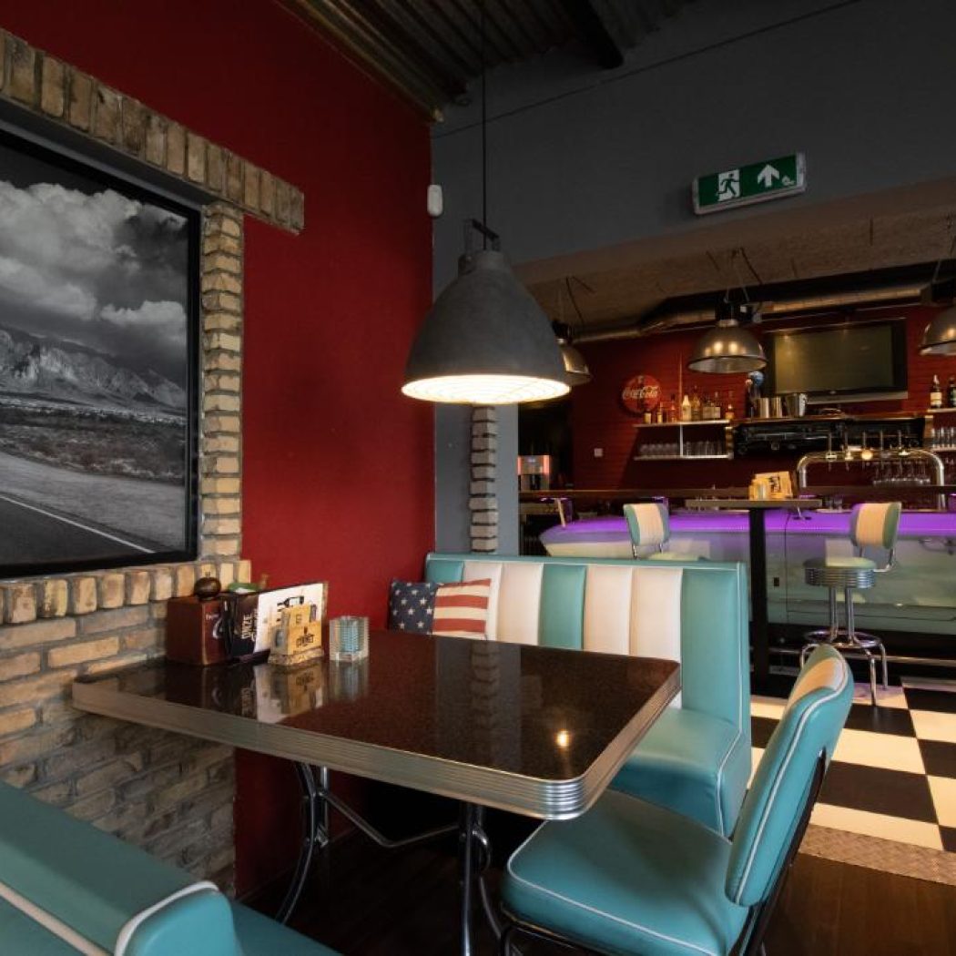 Amerikaans diner interieur met rode muren, turkoois-witte zithoek, zwart-wit landschapsfoto en bar met paarse verlichting.