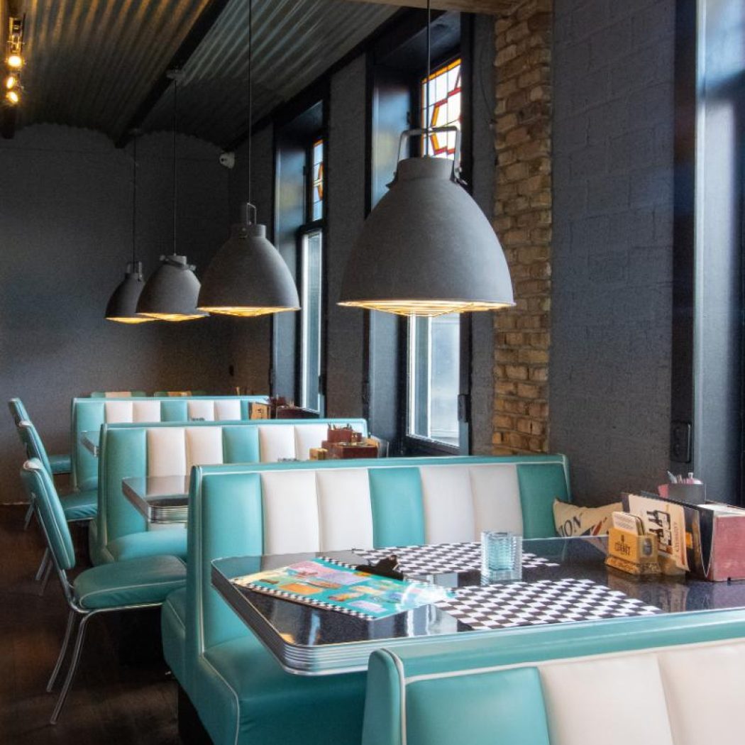 Retro diner met blauwgroene en witte banken, dambordtafels en grijze industriële lampen.